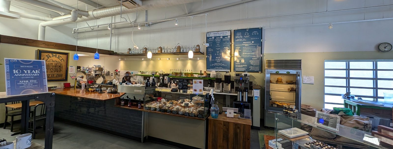 Inside Trifecta: the copper-topped coffee bar, menu boards, bakery case and chocolate counter under an open ceiling.