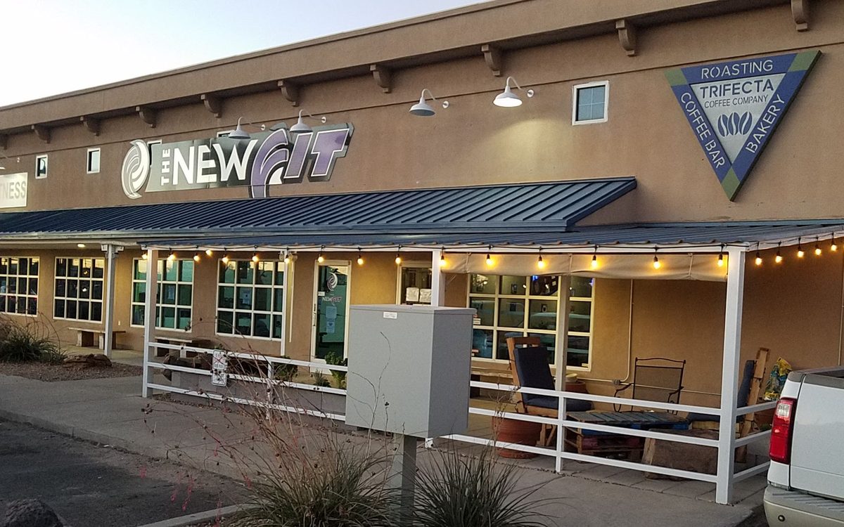 The Trifecta Coffee Company storefront on Montaño Rd at dusk: the triangle logo sign on a tan stucco building above a covered patio with string lights and outdoor seating.